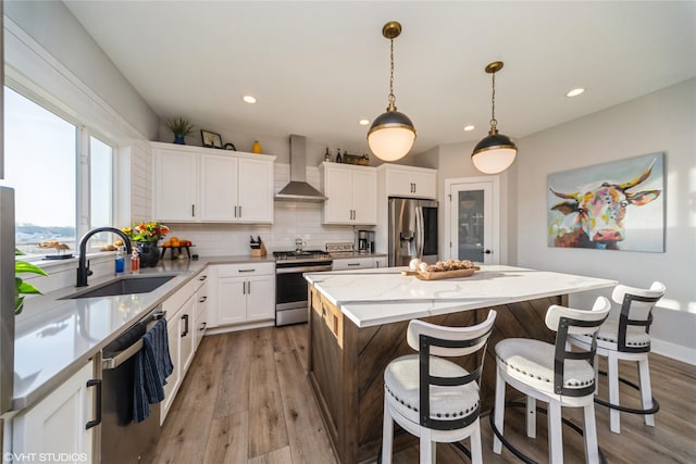 kitchen with light wood finished floors, a kitchen island, stainless steel appliances, wall chimney exhaust hood, and a sink