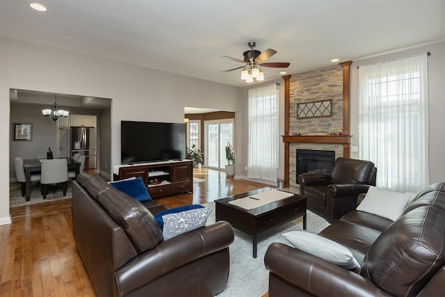 living area with a stone fireplace, ceiling fan with notable chandelier, light wood-type flooring, and baseboards