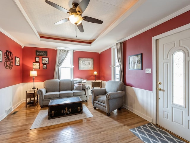 living room featuring a healthy amount of sunlight, ceiling fan, wainscoting, a raised ceiling, and light wood-type flooring