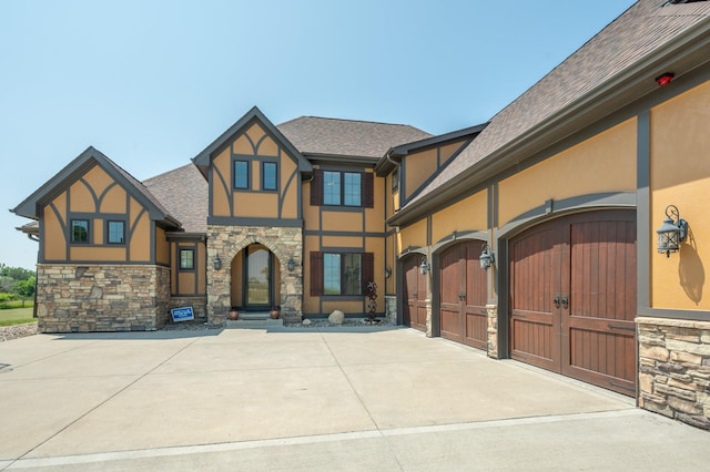 english style home with a shingled roof, stucco siding, concrete driveway, a garage, and stone siding