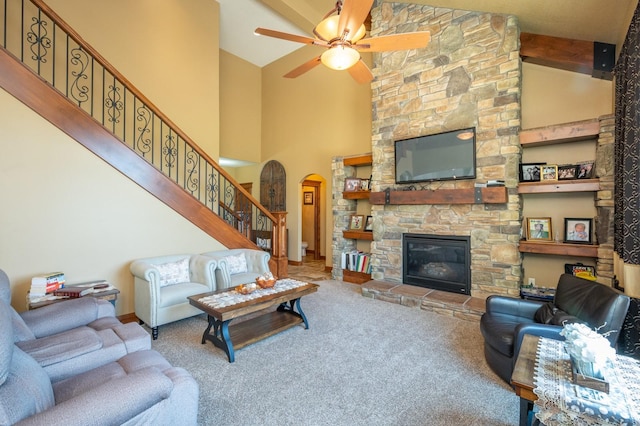 carpeted living area with beam ceiling, stairway, arched walkways, a stone fireplace, and ceiling fan