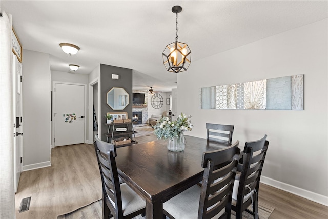 dining space with visible vents, light wood-style flooring, ceiling fan with notable chandelier, a stone fireplace, and baseboards