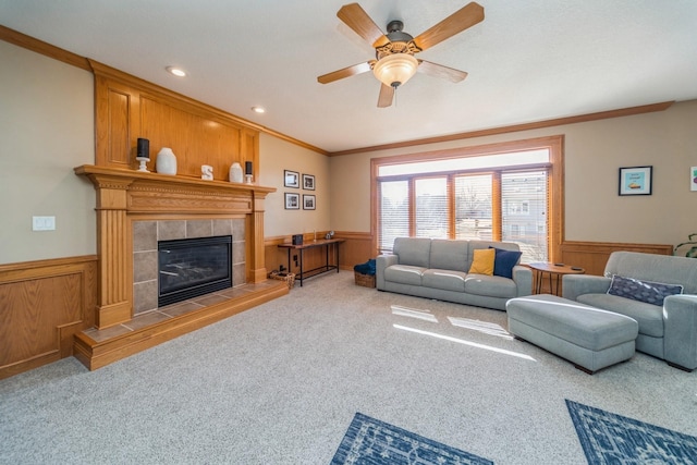 carpeted living area featuring wainscoting, a tile fireplace, ceiling fan, and ornamental molding