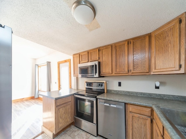 kitchen featuring light tile patterned floors, appliances with stainless steel finishes, a peninsula, brown cabinetry, and a textured ceiling