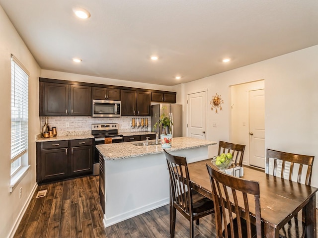 kitchen with an island with sink, appliances with stainless steel finishes, decorative backsplash, dark brown cabinets, and dark wood-style flooring