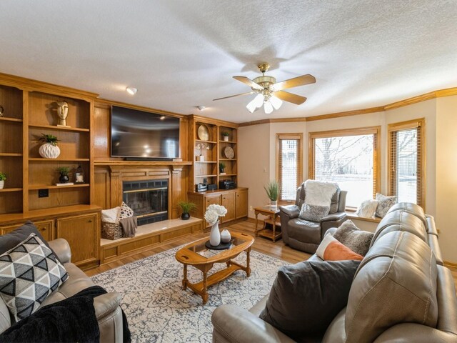 living room with light wood-style flooring, ornamental molding, a textured ceiling, a glass covered fireplace, and ceiling fan