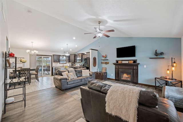 living area featuring vaulted ceiling, light wood-type flooring, ceiling fan with notable chandelier, baseboards, and a lit fireplace