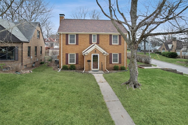 colonial home featuring brick siding, a front yard, roof with shingles, and a chimney