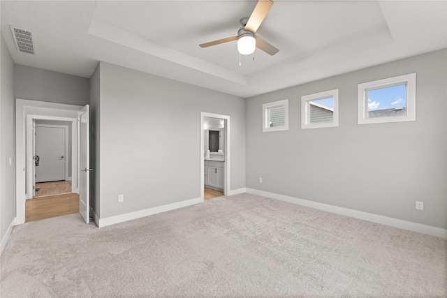 unfurnished bedroom featuring ensuite bathroom, a tray ceiling, light colored carpet, visible vents, and baseboards