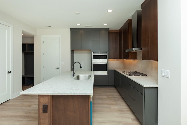 kitchen with wall chimney exhaust hood, recessed lighting, appliances with stainless steel finishes, a kitchen island with sink, and a sink