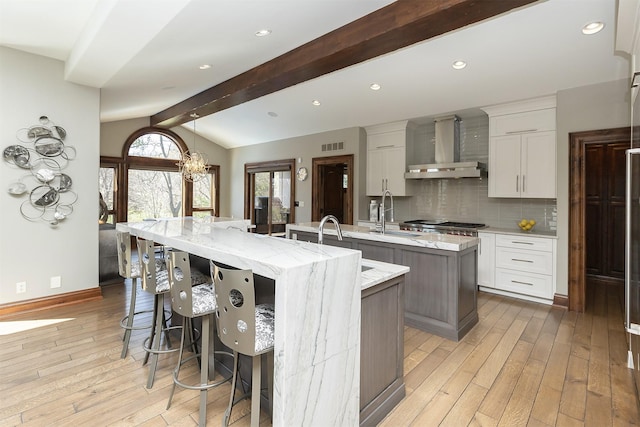kitchen with wall chimney range hood, a sink, light wood-style floors, vaulted ceiling with beams, and a large island