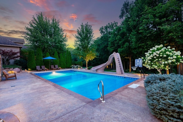 pool at dusk featuring a water slide, an outdoor pool, and a patio