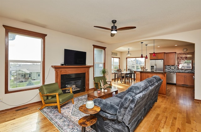 living room featuring light wood-style flooring, arched walkways, a glass covered fireplace, visible vents, and a ceiling fan