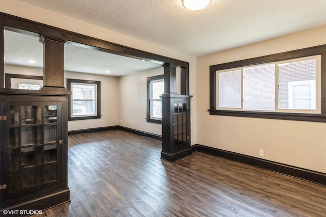 unfurnished living room featuring a textured ceiling, dark wood-style flooring, baseboards, and ornate columns