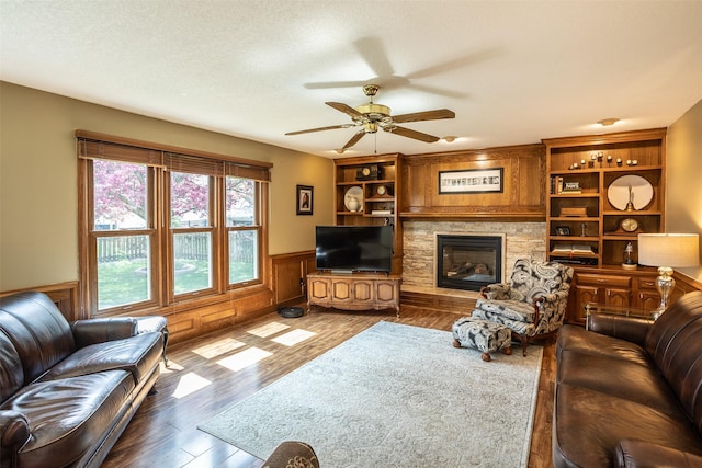 living area featuring a textured ceiling, a fireplace, wood finished floors, a ceiling fan, and wainscoting