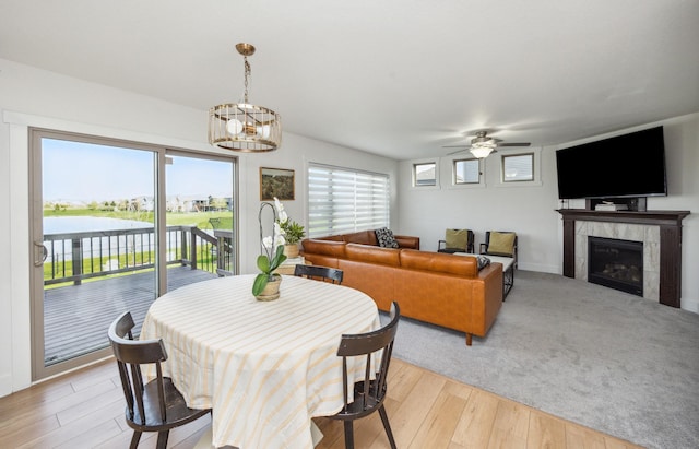 dining space with a tiled fireplace, light wood-style flooring, a water view, and ceiling fan with notable chandelier