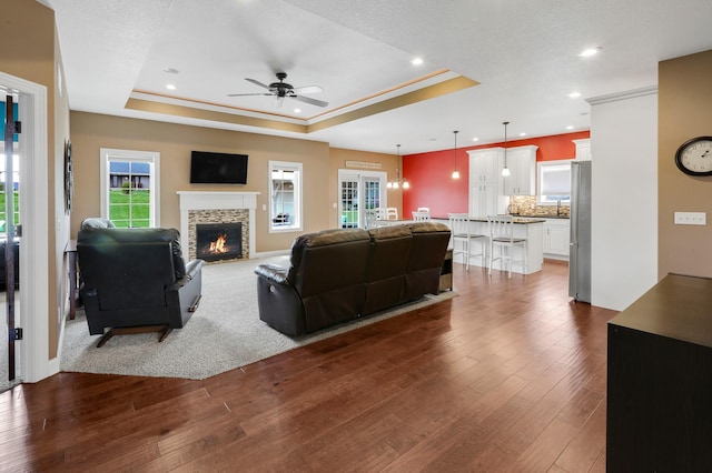 living area featuring a raised ceiling, ceiling fan, dark wood-style flooring, recessed lighting, and a fireplace