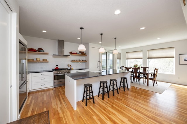 kitchen with open shelves, high end appliances, dark countertops, wall chimney range hood, and a kitchen bar