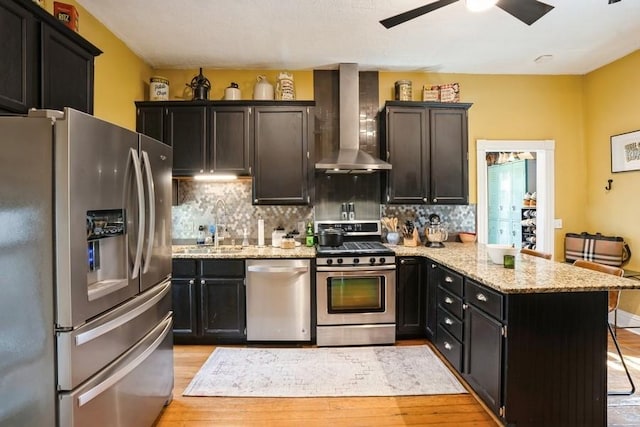 kitchen with a peninsula, wall chimney range hood, a sink, light wood finished floors, and stainless steel appliances