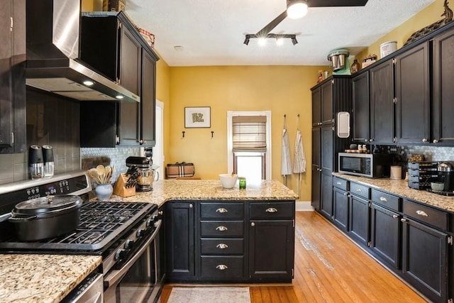 kitchen with wall chimney exhaust hood, stainless steel appliances, a ceiling fan, a peninsula, and decorative backsplash
