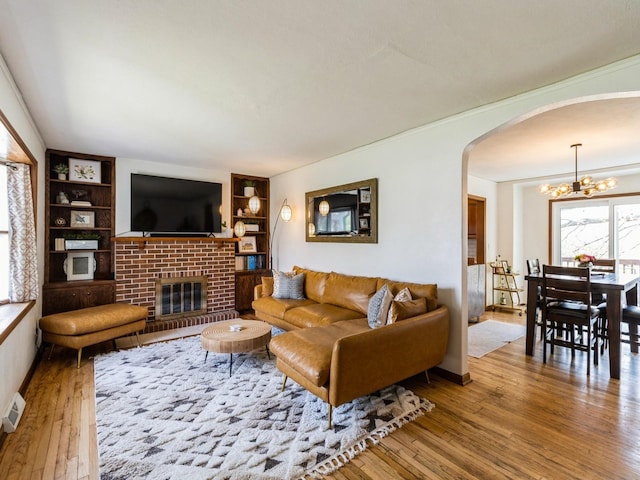 living area featuring wood-type flooring, built in features, a brick fireplace, a chandelier, and arched walkways
