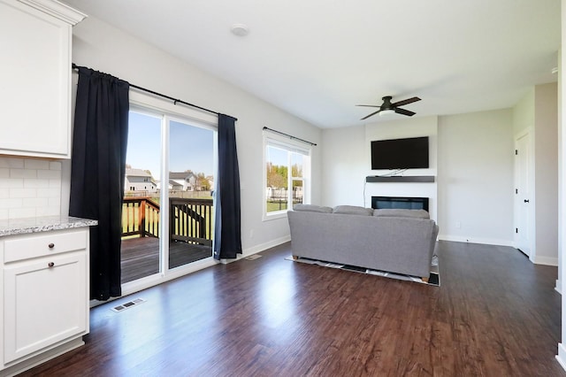 living area featuring visible vents, a glass covered fireplace, baseboards, ceiling fan, and dark wood-type flooring
