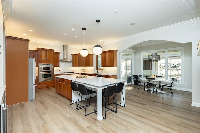 kitchen with arched walkways, appliances with stainless steel finishes, brown cabinetry, and wall chimney exhaust hood