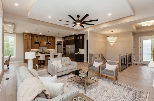 living room with a tray ceiling, baseboards, light wood finished floors, and ceiling fan with notable chandelier