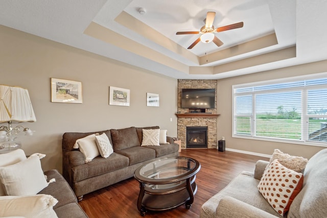 living area featuring ceiling fan, a stone fireplace, wood finished floors, baseboards, and a tray ceiling
