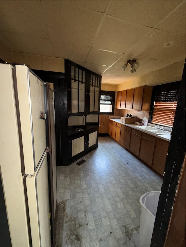 kitchen featuring brown cabinetry, visible vents, a sink, freestanding refrigerator, and a drop ceiling