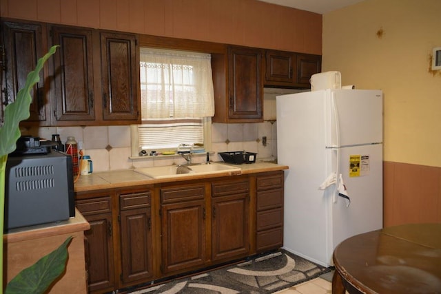 kitchen with a sink, freestanding refrigerator, tile countertops, dark brown cabinetry, and decorative backsplash
