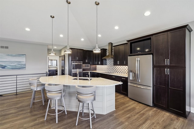 kitchen featuring wall chimney range hood, a breakfast bar, visible vents, a center island with sink, and appliances with stainless steel finishes