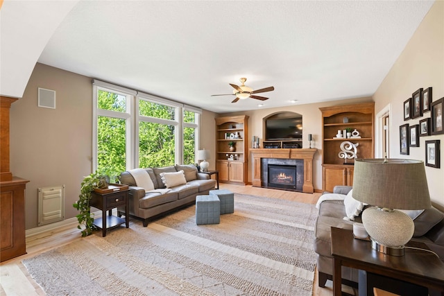 living area with a ceiling fan, light wood-style flooring, a glass covered fireplace, visible vents, and built in features