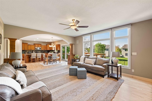 living area featuring ceiling fan with notable chandelier, ornate columns, light wood finished floors, arched walkways, and baseboards