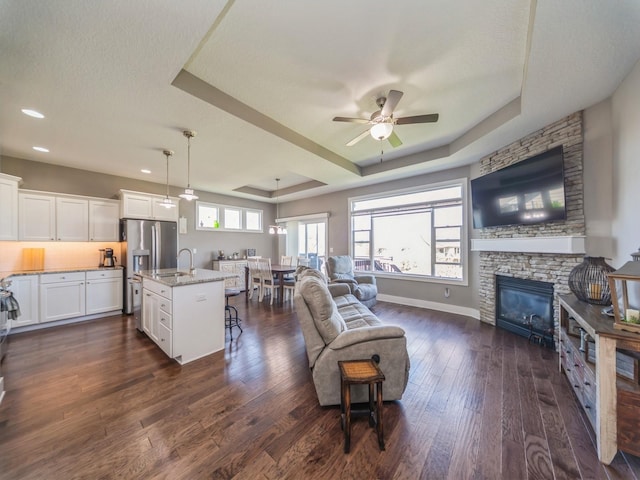 living area featuring a fireplace, dark wood-type flooring, baseboards, a raised ceiling, and ceiling fan