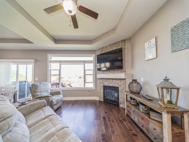 living area featuring a fireplace, dark wood-type flooring, baseboards, a raised ceiling, and ceiling fan