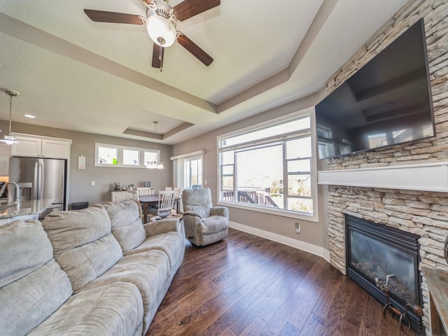 living room featuring a tray ceiling, a fireplace, dark wood-style flooring, baseboards, and ceiling fan