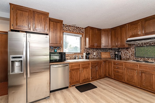 kitchen featuring appliances with stainless steel finishes, a sink, under cabinet range hood, a textured ceiling, and light wood finished floors