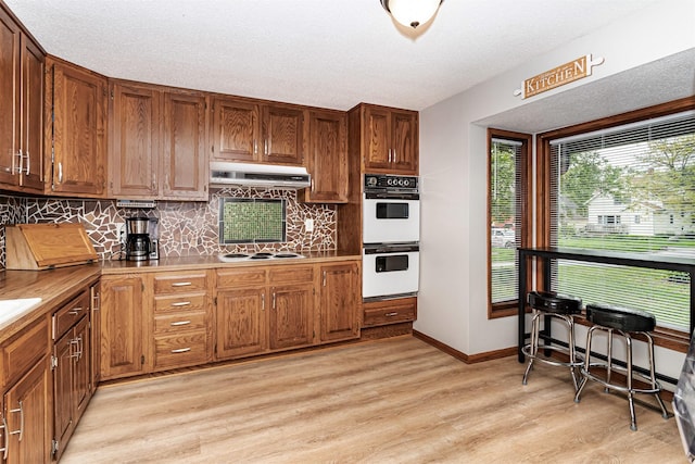 kitchen with white appliances, under cabinet range hood, light wood-type flooring, decorative backsplash, and light countertops