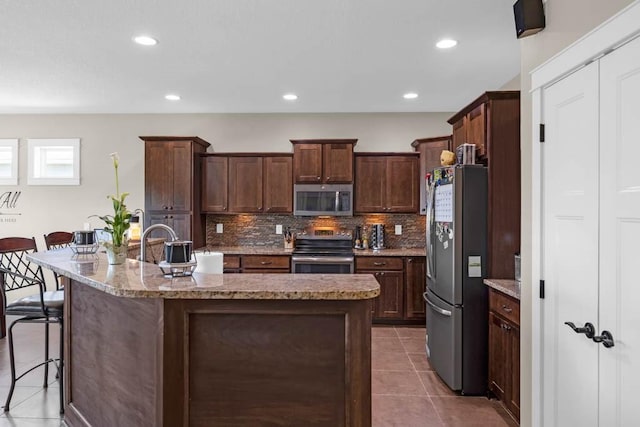 312 Evergreen Dr NW, Bondurant, IA 50035 - kitchen featuring appliances with stainless steel finishes, decorative backsplash, light tile patterned floors, a kitchen island with sink, and a kitchen bar