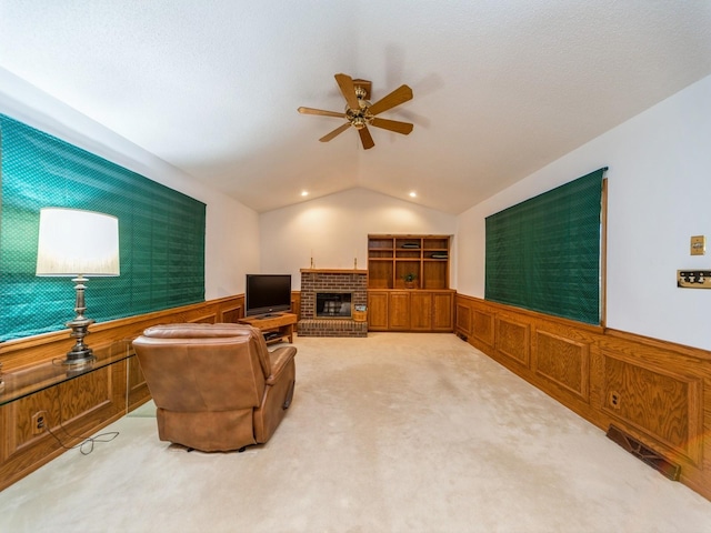 carpeted living room with ceiling fan, a wainscoted wall, lofted ceiling, and a brick fireplace