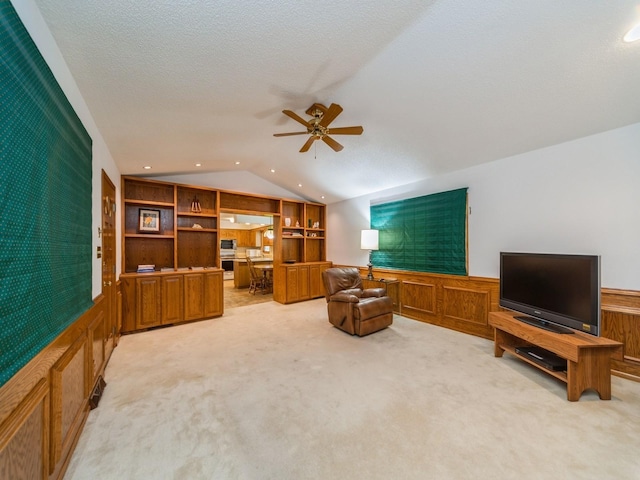 living area with lofted ceiling, ceiling fan, light carpet, a wainscoted wall, and built in shelves
