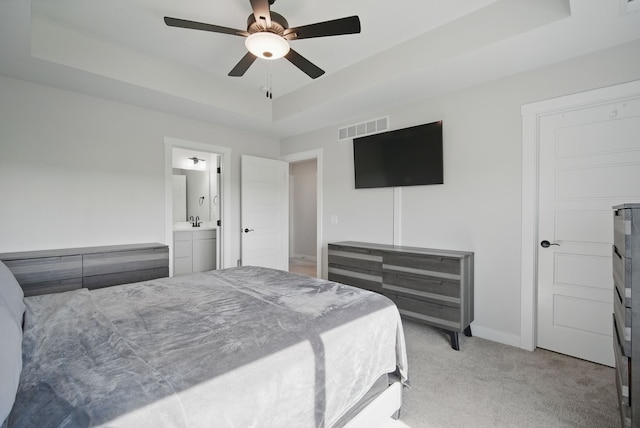 bedroom featuring a tray ceiling, light carpet, baseboards, ensuite bath, and a ceiling fan