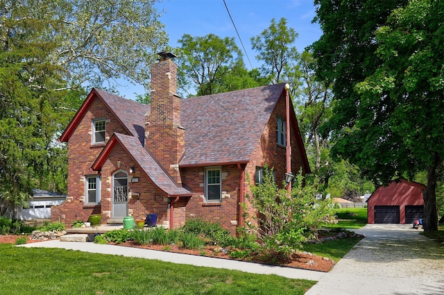 tudor-style house featuring an outbuilding, a chimney, a detached garage, brick siding, and a front yard