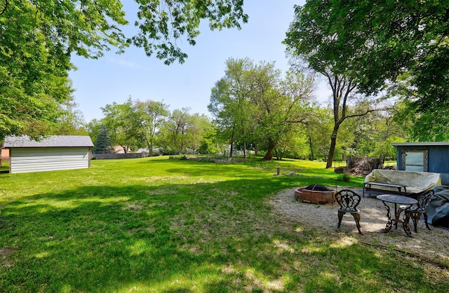 view of grassy yard featuring an outdoor fire pit, view of wooded area, and an outbuilding