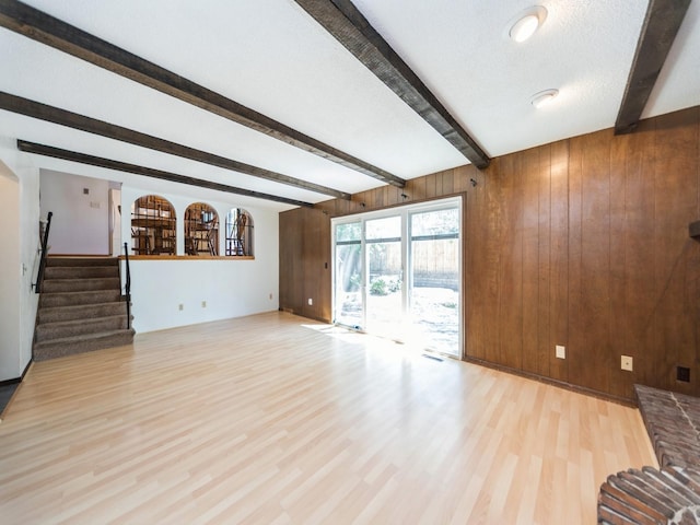 unfurnished living room featuring beam ceiling, wood finished floors, wooden walls, stairway, and baseboards