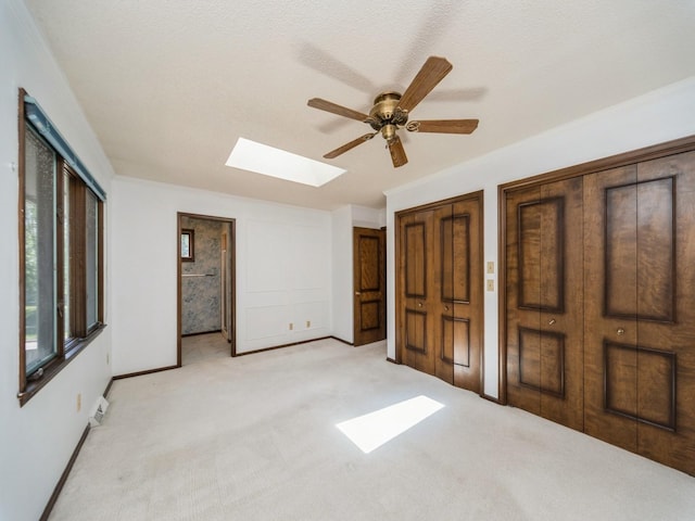 unfurnished bedroom with a skylight, light carpet, multiple closets, baseboards, and a ceiling fan