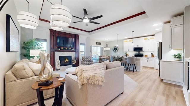 living room featuring ceiling fan, a raised ceiling, recessed lighting, light wood-style flooring, and a glass covered fireplace