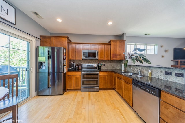 kitchen with appliances with stainless steel finishes, a sink, brown cabinetry, dark stone counters, and light wood-type flooring