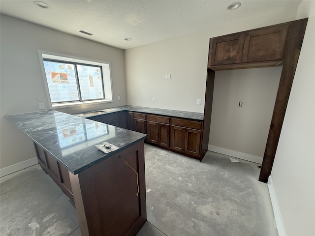 kitchen featuring a peninsula, a textured ceiling, dark brown cabinets, concrete floors, and dark stone countertops